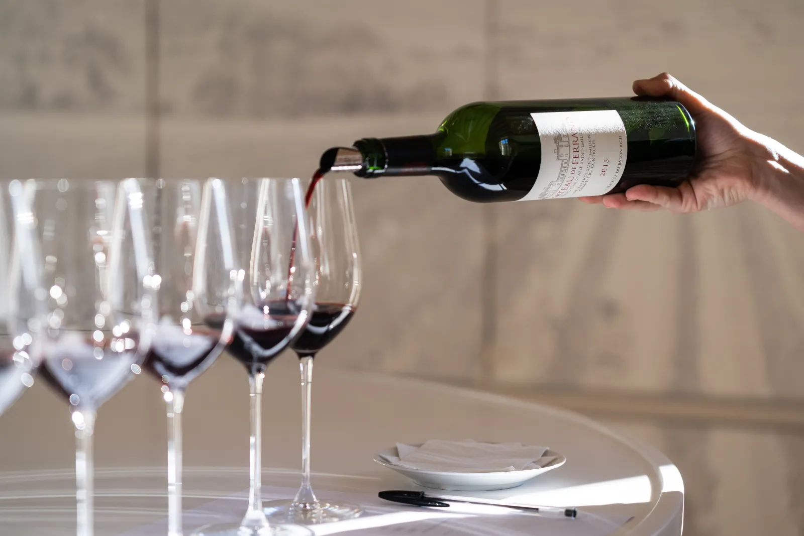 A hand pouring red wine from a Bordeaux bottle into lined tasting glasses on a table, with a white plate and cutlery in the background.