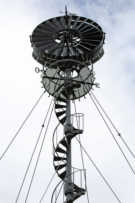 Bollée wind turbine at Beauval Estate in Bordeaux, low-angle view showing the metal structure with black helical blades, central mast and tension cables against a cloudy sky.
