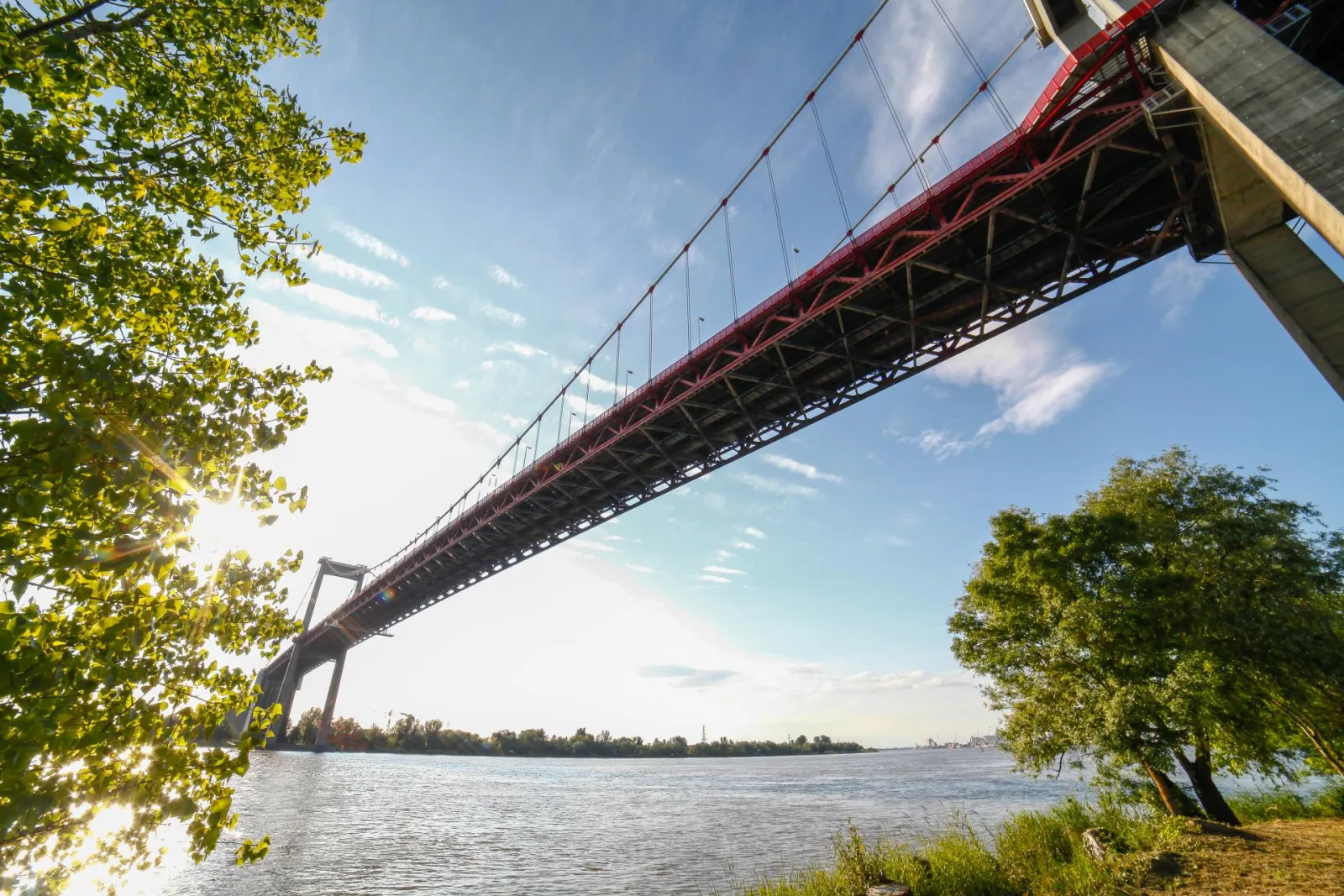 The Aquitaine Bridge, a large red and white suspension bridge spanning the Garonne River in Bordeaux, framed by green riverbank vegetation.