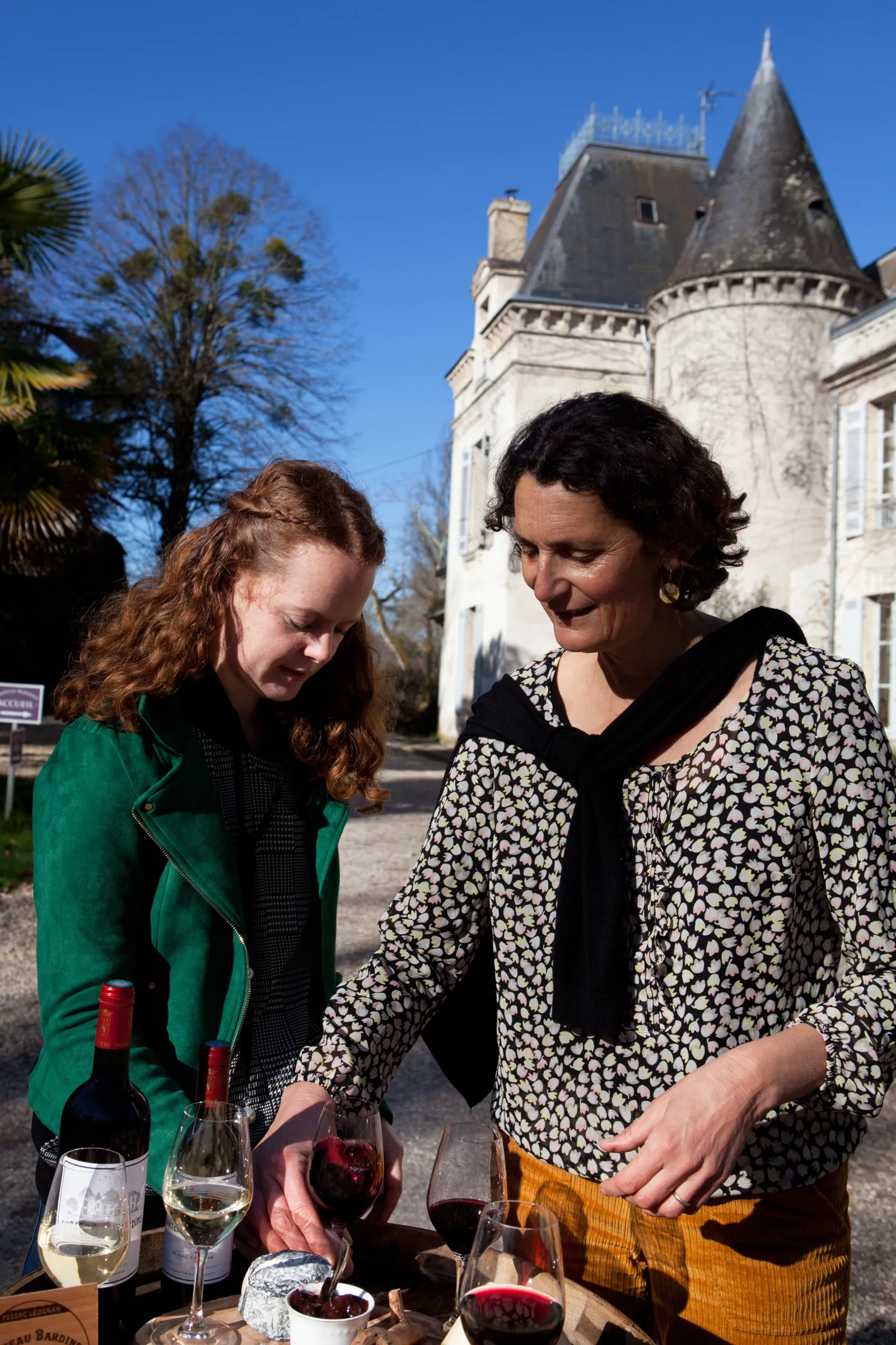 Two women enjoying wine tasting in front of a castle with glasses and bottles on a table, at the Château Workshops in Bordeaux.