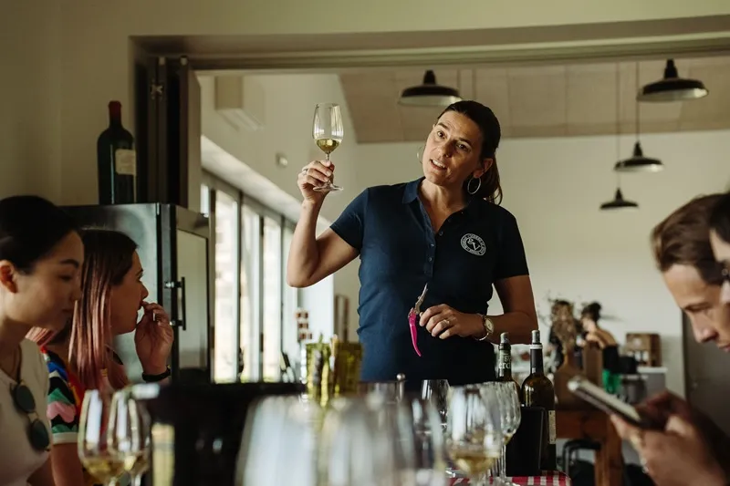 A sommelier presents a glass of white wine to visitors during a tasting at Château Le Grand Verdus in Bordeaux, in a modern room with pendant lights.