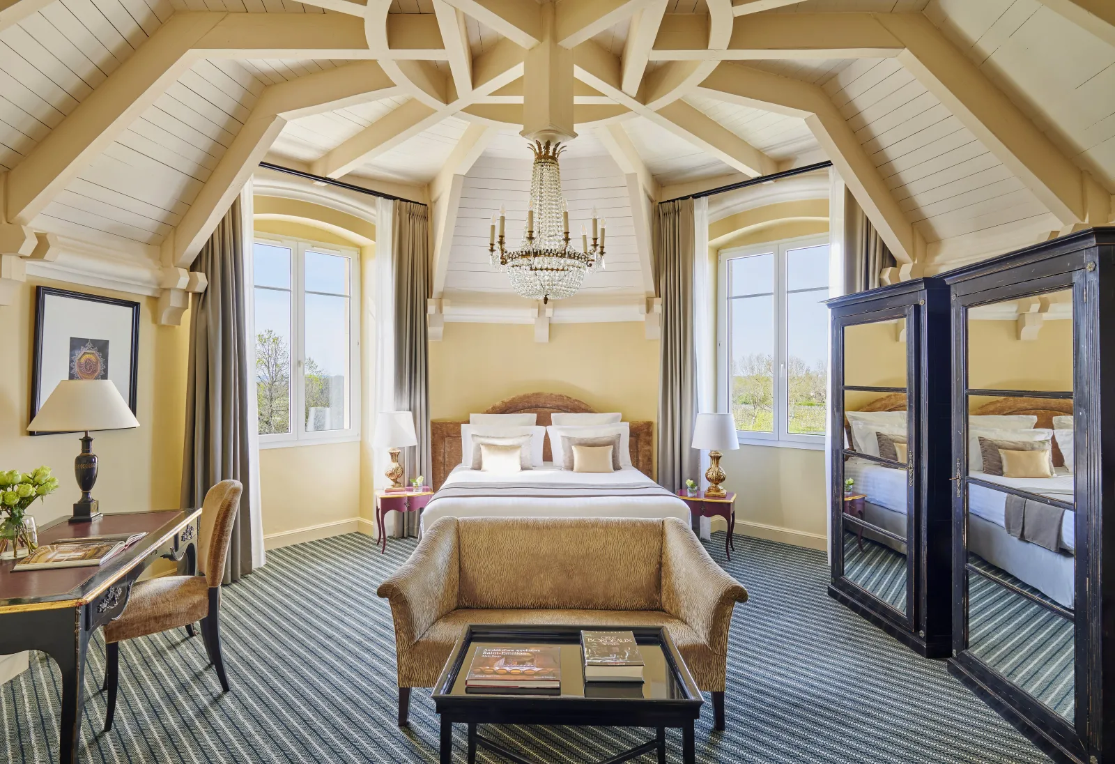 Luxurious suite at Château Hôtel Grand Barrail in Bordeaux featuring vaulted white ceiling, crystal chandelier, king-size bed, elegant furnishings and large windows overlooking the vineyard