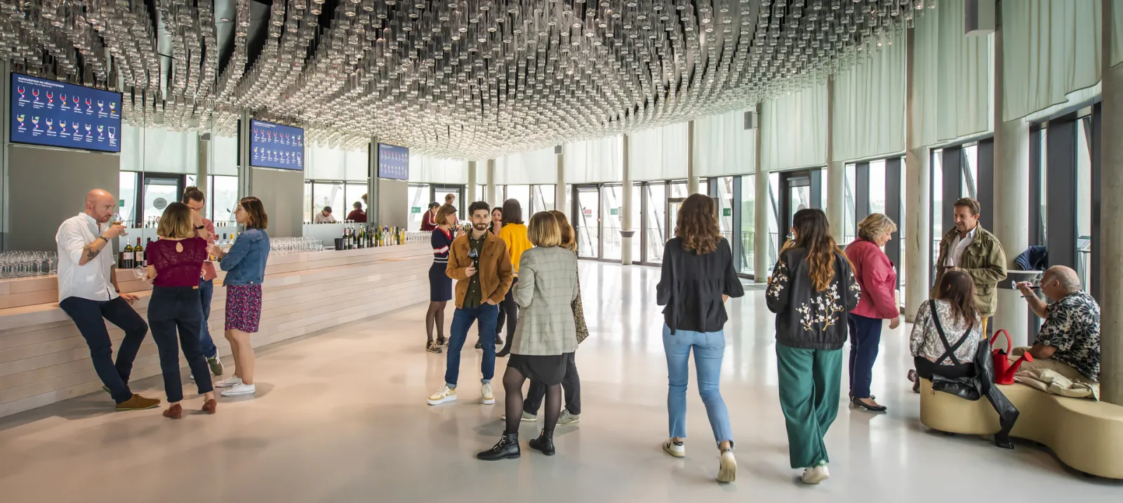 Modern interior of the Cité du Vin in Bordeaux featuring a geometric white ceiling, scattered visitors discussing and wine tasting, panoramic windows, and a tasting bar on the left.