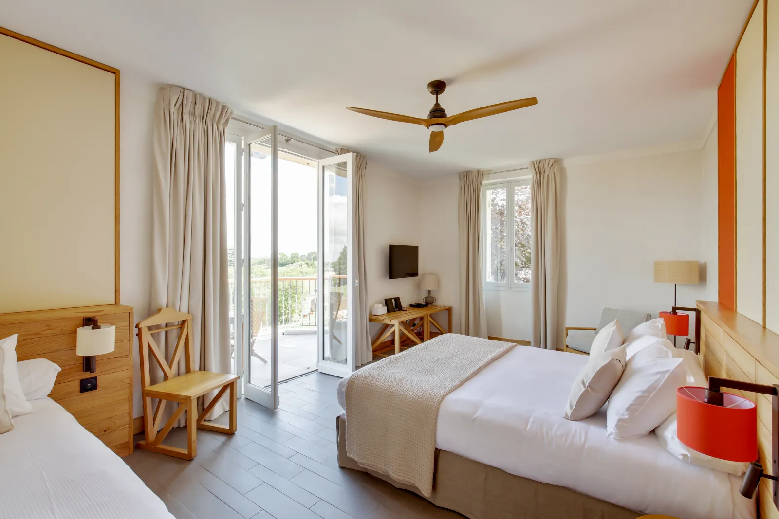 Spacious hotel bedroom featuring two beds, light wood furniture, orange accents, and large glass doors opening onto a balcony overlooking Bordeaux vineyards