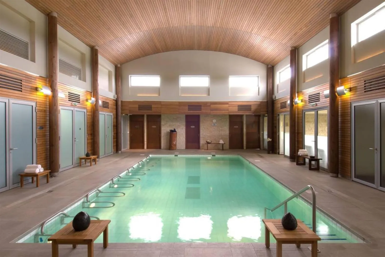 Indoor heated swimming pool at Hôtel Le Relais de Margaux in Bordeaux, featuring an Olympic-length pool, wooden changing rooms, arched wooden ceiling, and natural lighting from windows.
