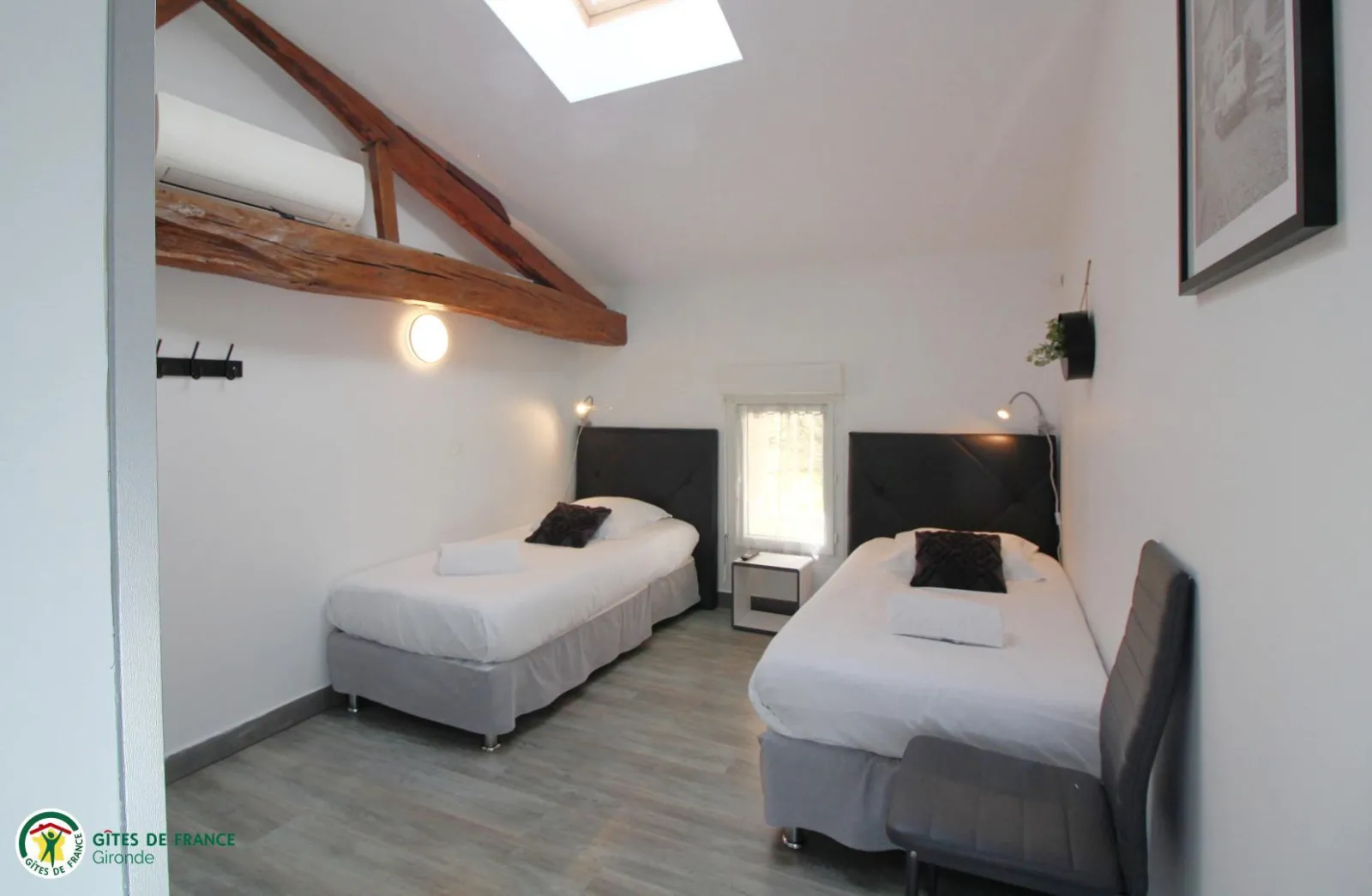 Guest room featuring two single beds with black headboards, white linens and black pillows, beneath an exposed wooden beam, with skylight and minimalist furnishings.