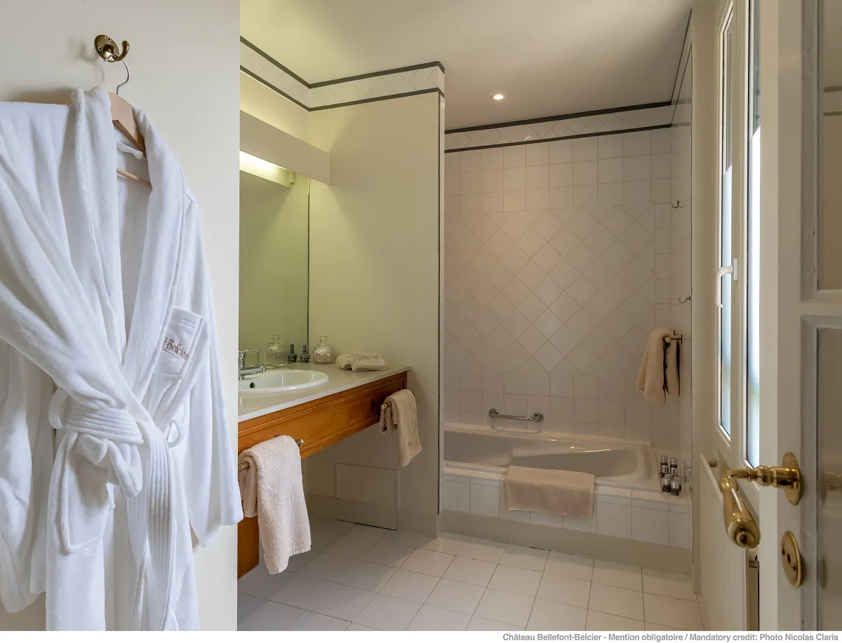 Elegant bathroom at Château Bellefont-Belcier in Bordeaux, featuring a wooden vanity, mirror, tiled bathtub, and white bathrobe