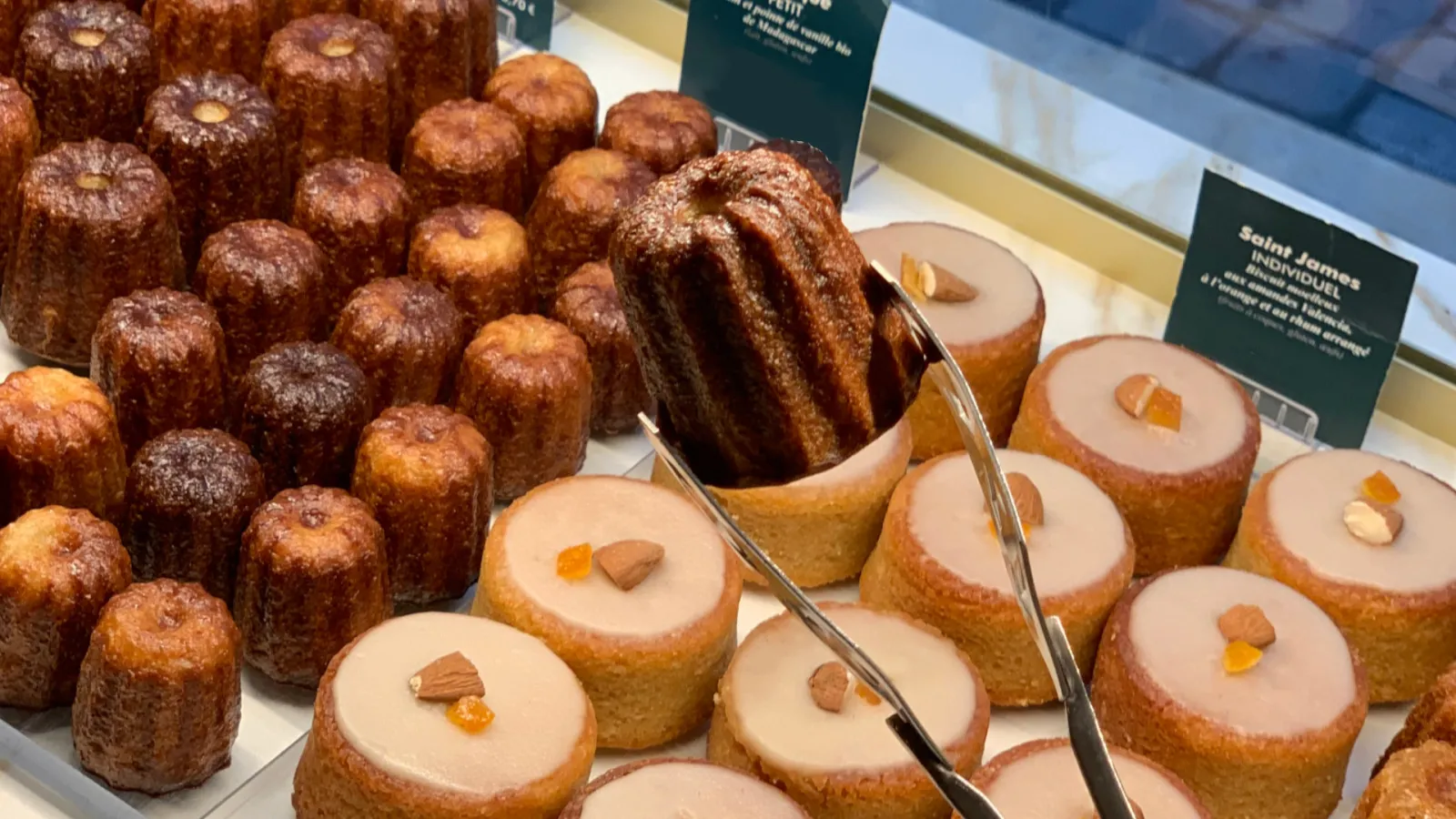 Bakery display case featuring Bordeaux cannelés and Saint-James pastries with icing, decorated with candied fruits and almonds