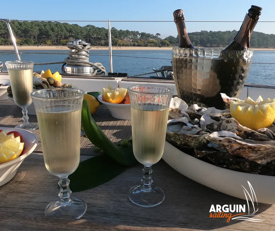 Tasting table aboard a sailing yacht with champagne glasses, fresh oysters, lemons and champagne bottles, overlooking the Gironde estuary and wooded coastline.