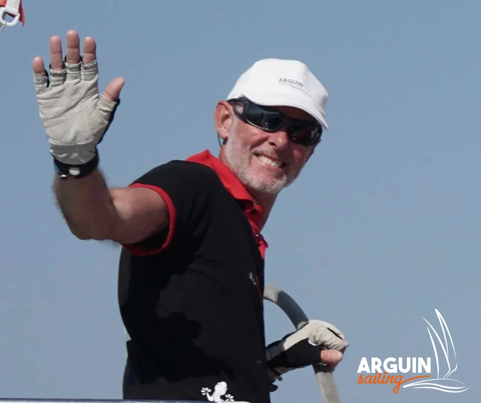 Smiling man on a sailboat wearing a white Arguin cap, sailing gloves and holding a sail, waving with the Arguin Sailing logo visible in the background.