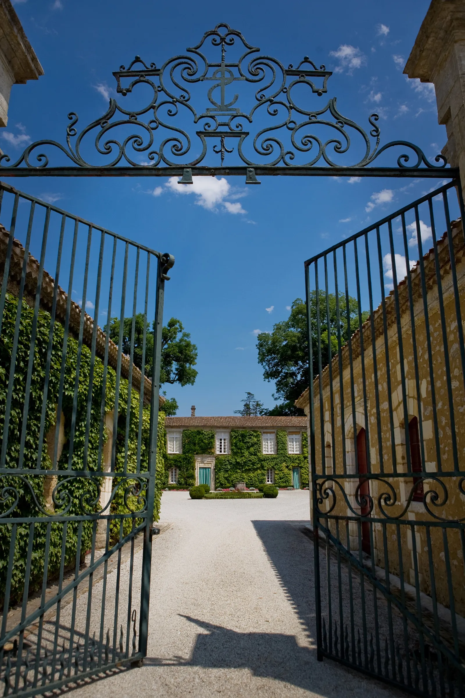 View through ornamental iron gates of Château Carbonnieux in Bordeaux, showing the courtyard with the main building with green shutters, surrounded by ivy and clear blue sky.