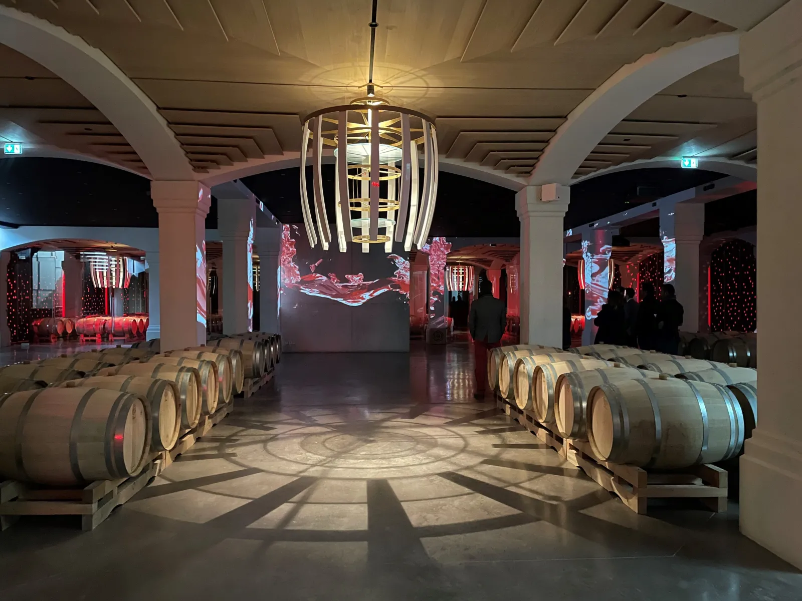 Interior of Château Montlabert wine cellar in Bordeaux featuring rows of wooden barrels, red ambient lighting, central chandelier and architectural columns