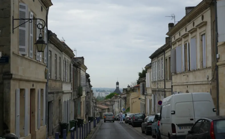 Charming street in Old Lormont, Bordeaux, lined with classical stone buildings in ochre and white, overlooking the city skyline with parked cars along the road.