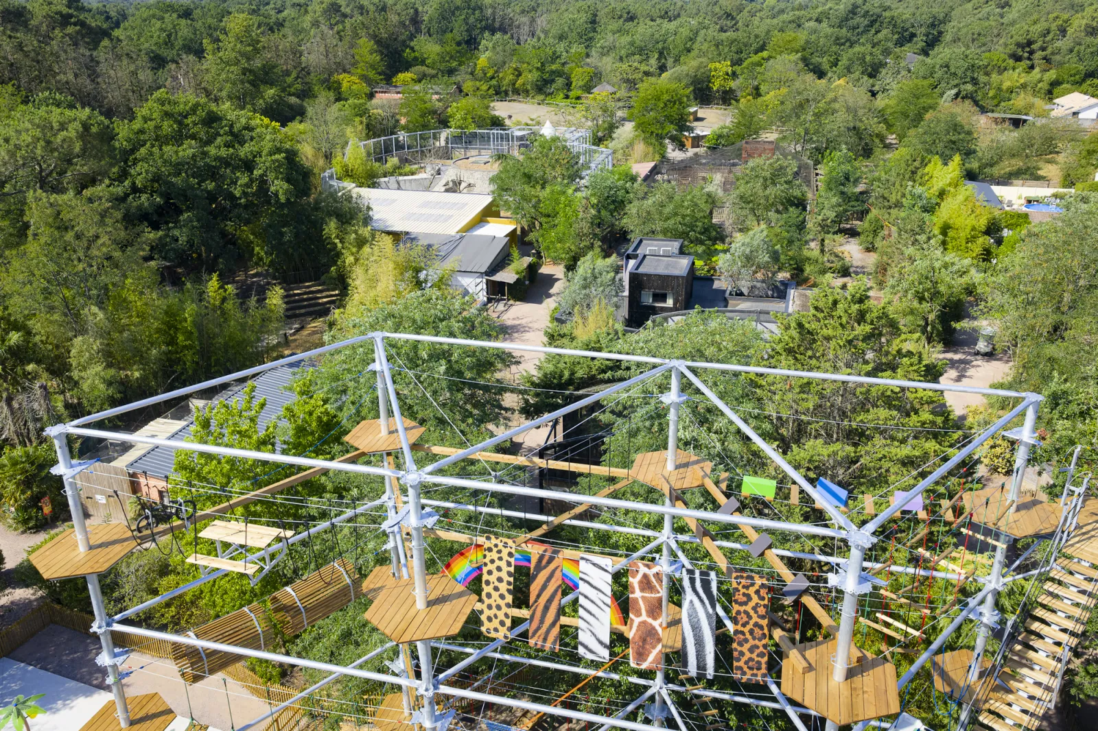 Aerial view of an observation platform with white metal structure and wooden deck overlooking a dense forest with scattered buildings and recreational facilities at Les Cimes de Bordeaux Pessac.