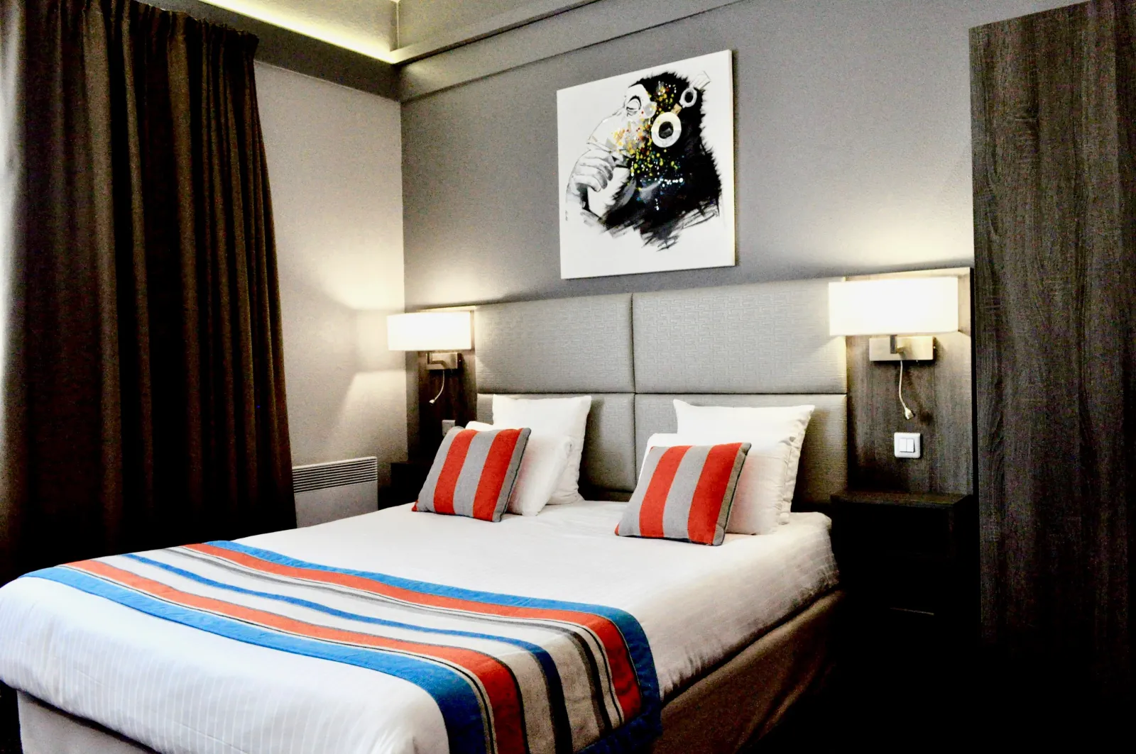 Modern hotel room featuring a double bed with white linens and blue-red striped bedding, red striped pillow, grey upholstered headboard, side lamps, brown curtains, and monkey artwork with headphones on the wall