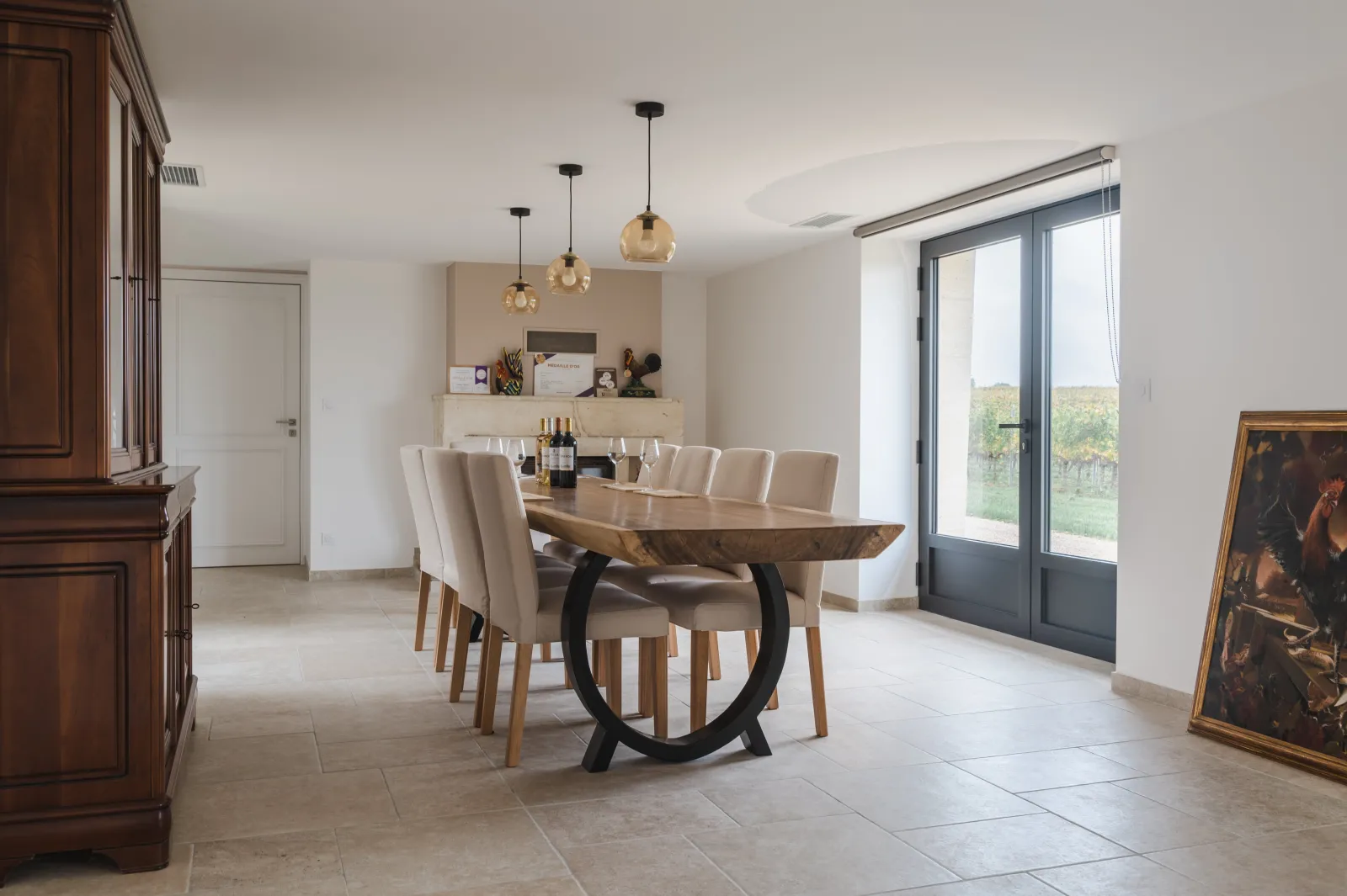 Elegant dining room at Château l'Eperon in Bordeaux featuring a massive wooden table with black legs, beige chairs, golden pendant lights, glass doors overlooking vineyards, and natural wood furniture.
