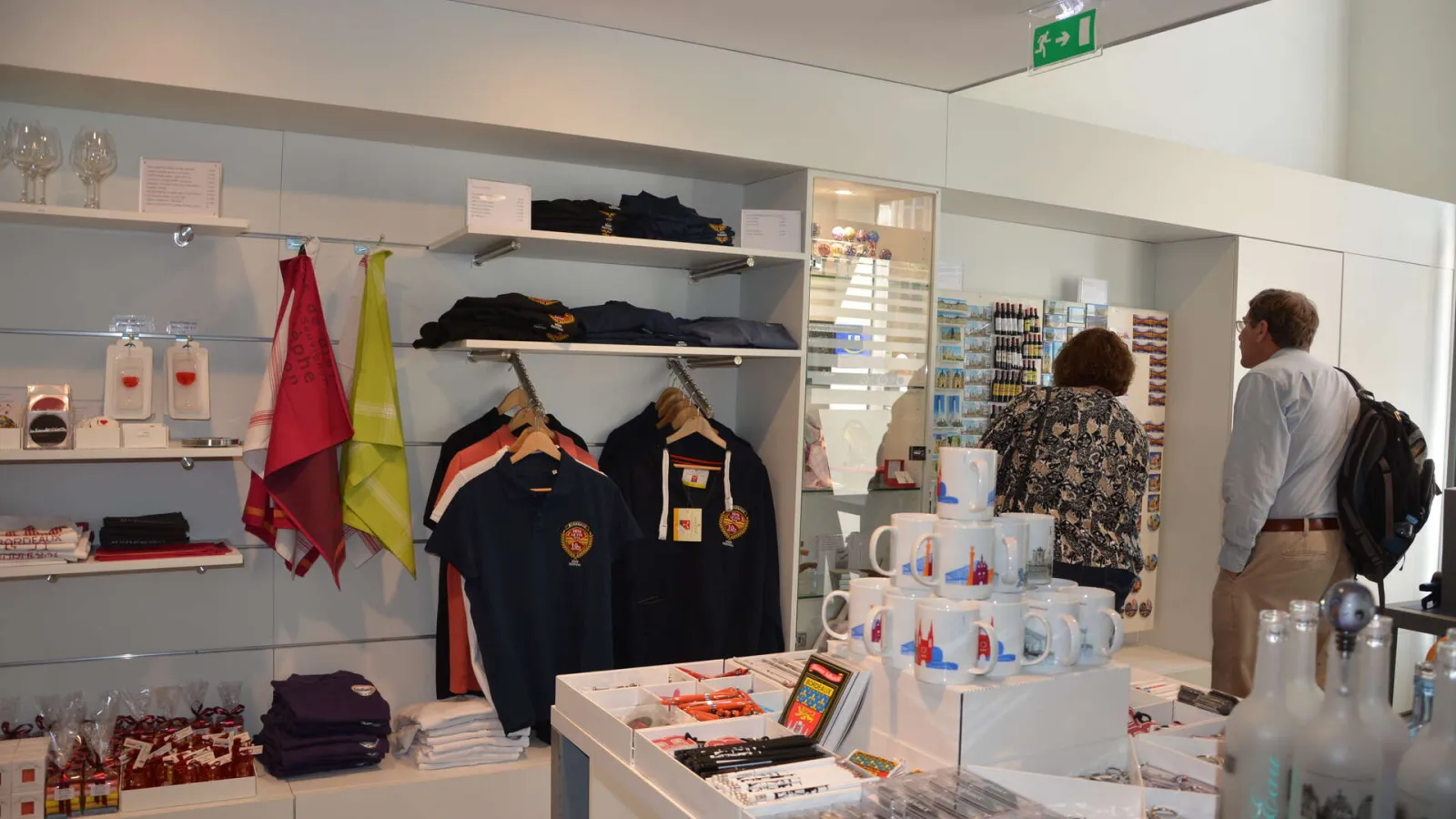 Interior of the Bordeaux Tourism Office shop displaying clothing, regional flags, mugs and souvenir products on shelves, with two visitors browsing merchandise