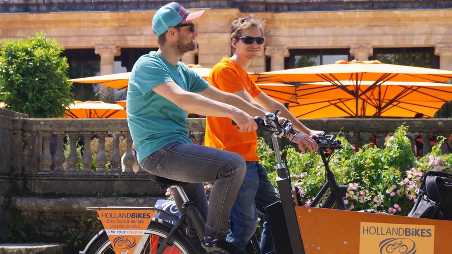 Two people riding an orange Holland Bikes rental bicycle in Bordeaux, with orange umbrellas and flowers in the background