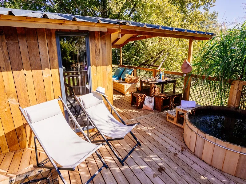 Wooden deck terrace of an eco-friendly accommodation featuring sun loungers, yellow cabin, pergola, round hot tub and lush green surroundings at EcÔtelia Domain in Bordeaux.