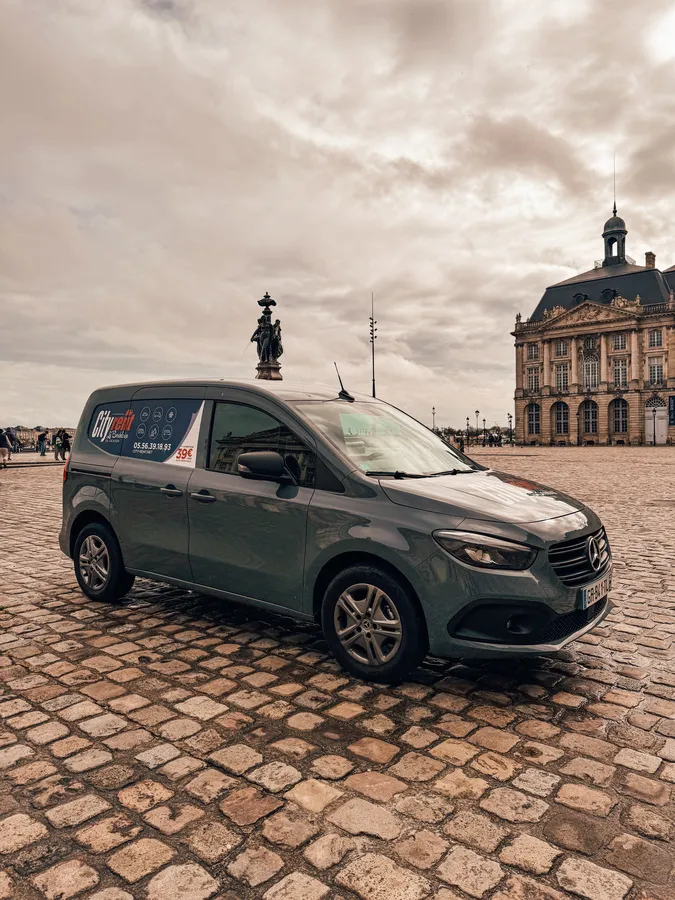 A gray-blue rental Mercedes minibus parked on Place de la Bourse in Bordeaux, with the Stock Exchange building in the background and an equestrian statue in the center of the square