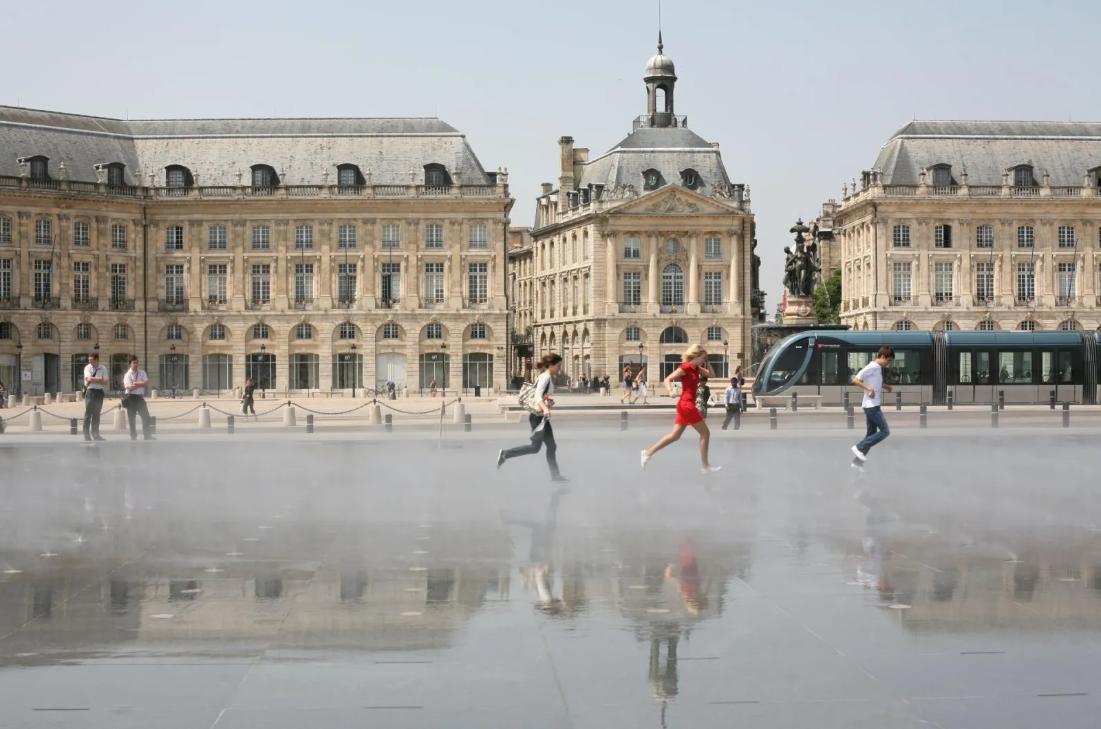 Bordeaux's Place de la Bourse featuring classical golden stone buildings, a reflective water mirror plaza, people jogging and walking, and a modern tram passing through the square.