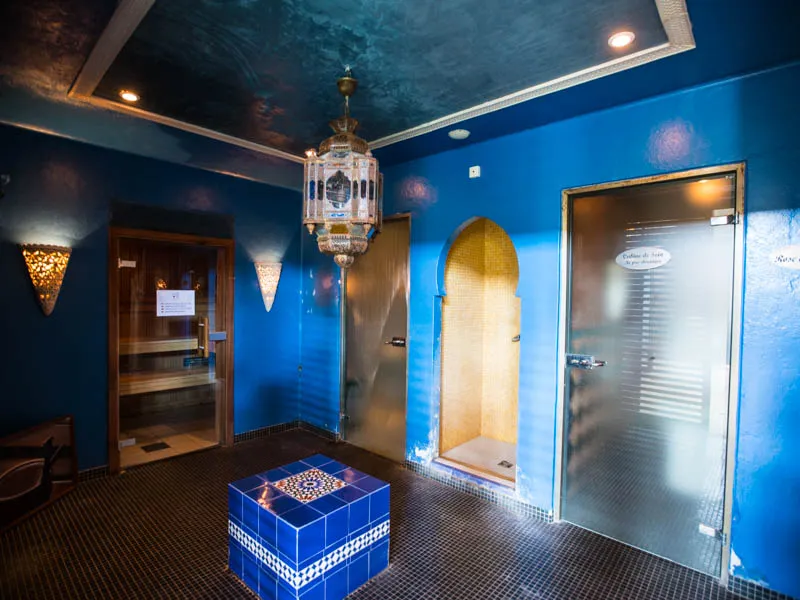 Interior of Les Bains de Margaux spa in Bordeaux featuring vibrant blue walls, textured ceiling, Moroccan chandelier, golden treatment cabin doors, and central pool with blue zellige tiles.