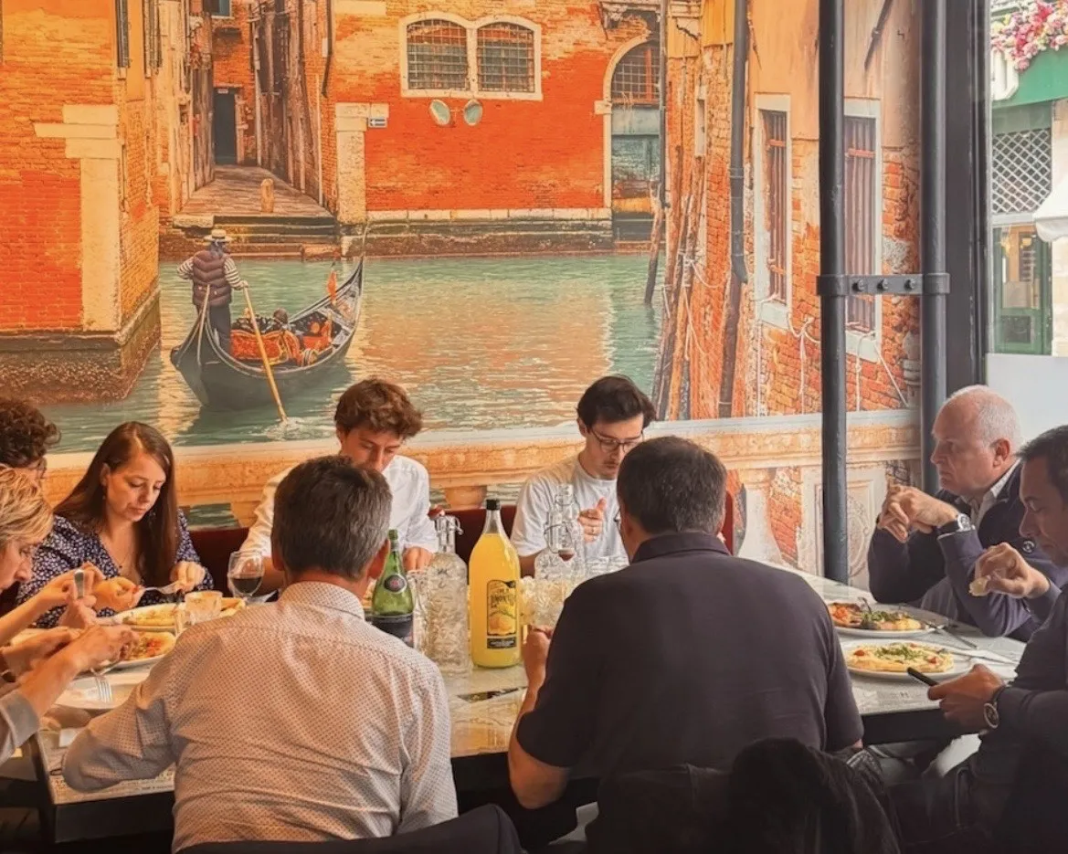 Group of people dining in a restaurant overlooking a Venetian canal with red brick buildings and a gondola in the background.