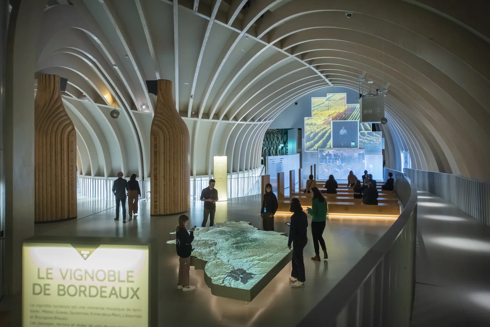 Interior of the Cité du Vin in Bordeaux featuring a modern arched white ceiling design, visitors viewing an illuminated scale model of the Bordeaux vineyard, and exhibition screens on the walls.