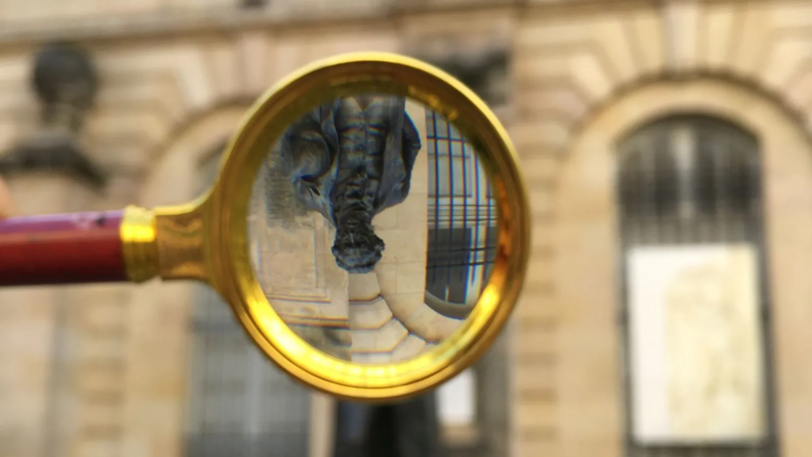 View through a golden magnifying glass of a bronze statue in Bordeaux with blurred historic buildings in the background