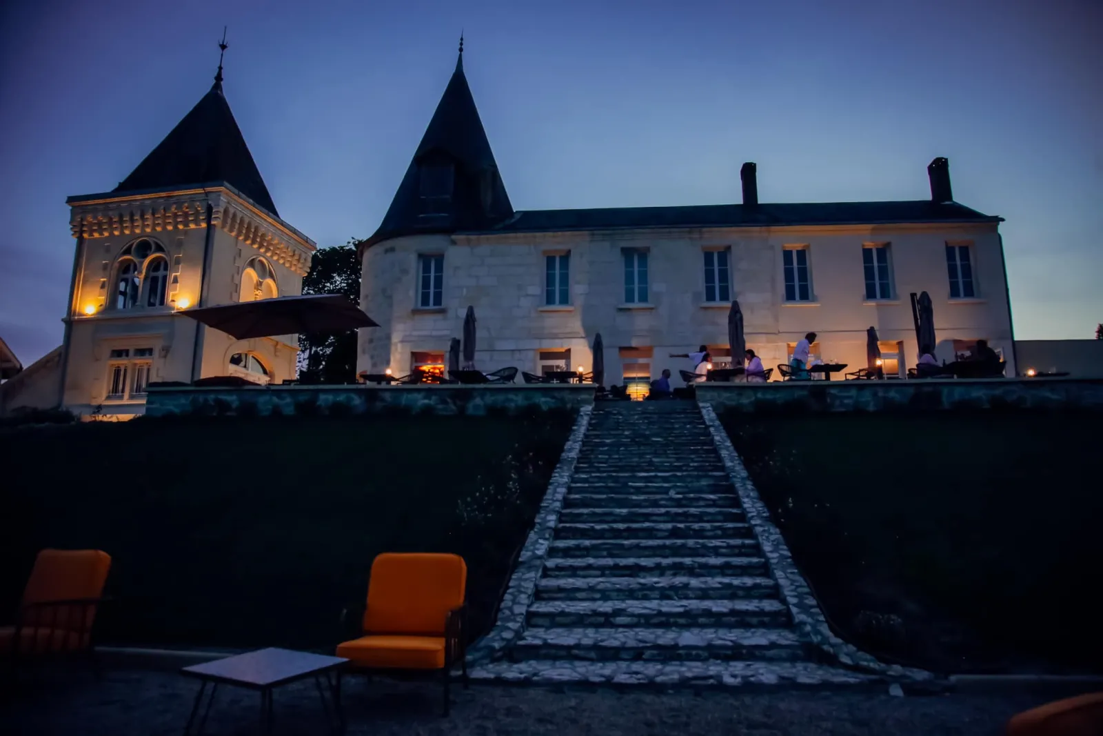 Illuminated castle at dusk featuring a pointed tower on the left, a stone pathway leading to the main façade, and orange chairs in the foreground in Bordeaux.