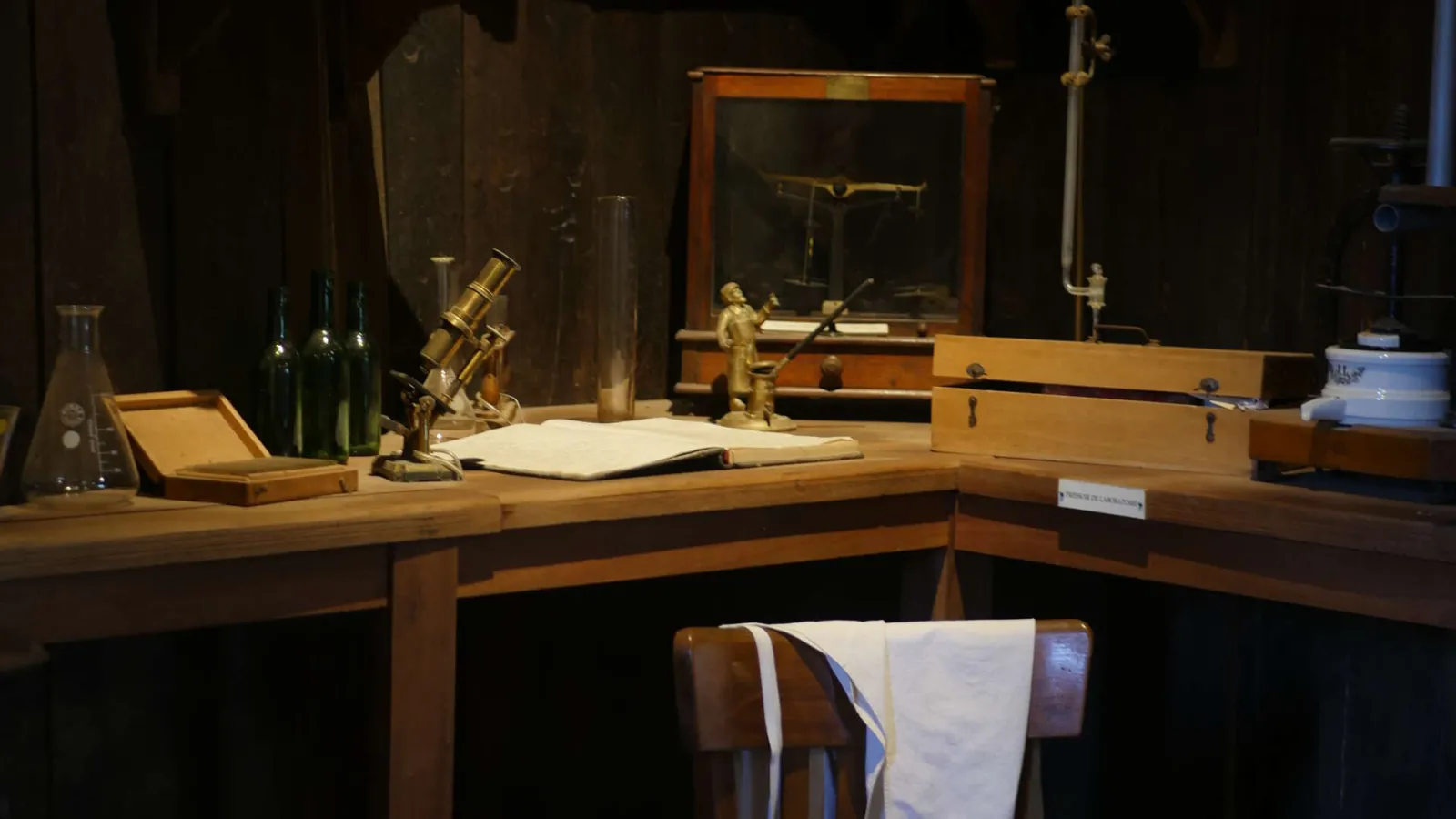 19th-century scientist's desk recreation featuring a brass microscope, wine bottles, books, measuring instruments and wooden furniture displayed in a museum display case.