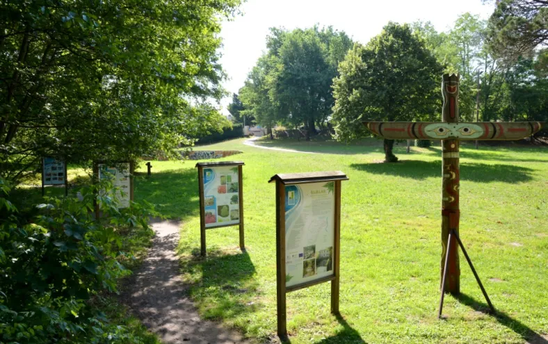 Information boards and educational panels along a path in a lush park at Moulin Bidon site in Bordeaux, with trees and a wooden cross visible.