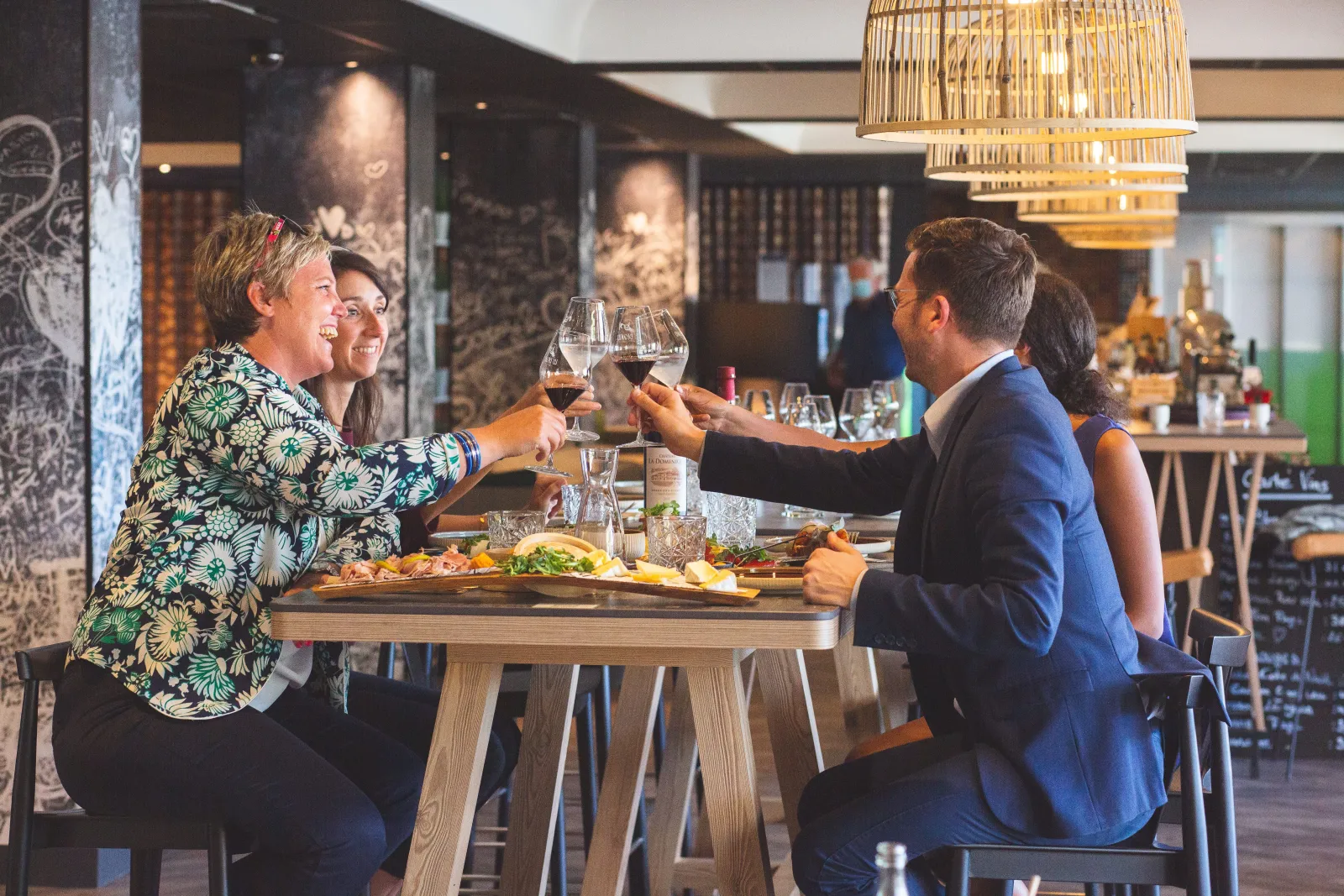 Four people in smart clothing toast with glasses of red wine around a wooden table in a modern Bordeaux restaurant, with dishes and water glasses in front of them.