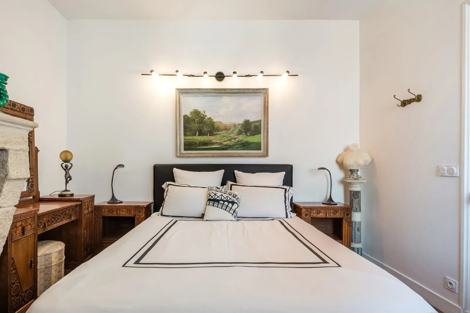Elegant bedroom featuring a black bed with white bordered linens, ornate wooden nightstands on both sides, landscape painting above the headboard, and brass wall sconces.