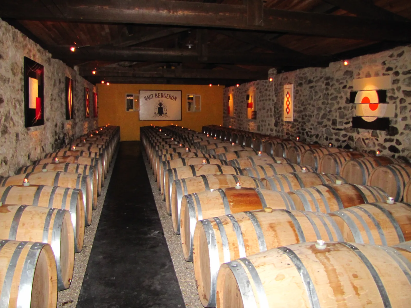 Aging cellar at Château Haut-Bergeron in Bordeaux featuring symmetrical rows of oak barrels lit by warm lighting and candles, with exposed stone walls.