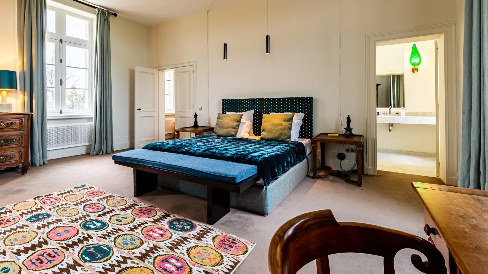 Spacious bedroom at Château Malescasse in Bordeaux featuring a king-size bed in blue and gold tones, ensuite bathroom, dark wood furniture, geometric patterned rug, large windows with grey-green curtains, and elegant décor.