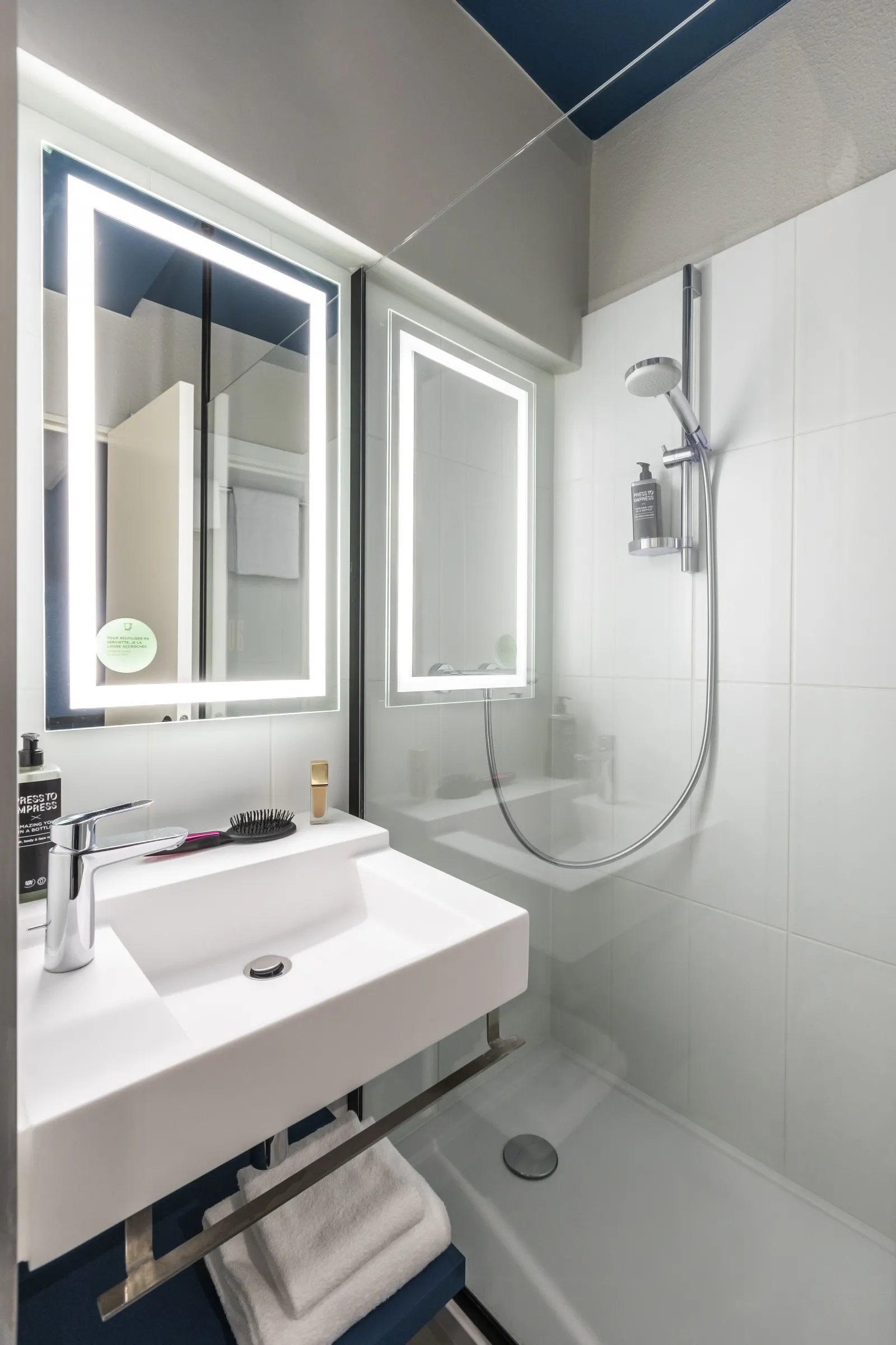 Modern bathroom at Ibis Styles Bordeaux Meriadeck featuring a white sink, illuminated mirror, walk-in shower with white tiles, and contemporary blue and grey ceiling
