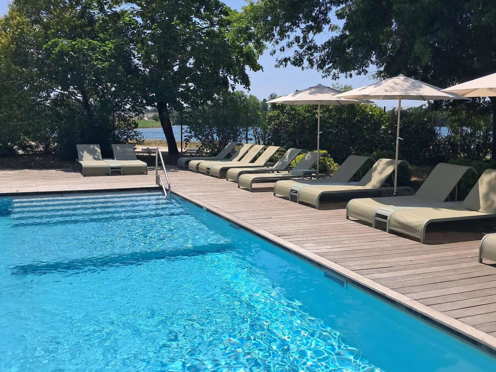 Outdoor swimming pool at Novotel Bordeaux Lac featuring turquoise water, wooden deck, beige loungers with umbrellas, and lake view surrounded by trees.