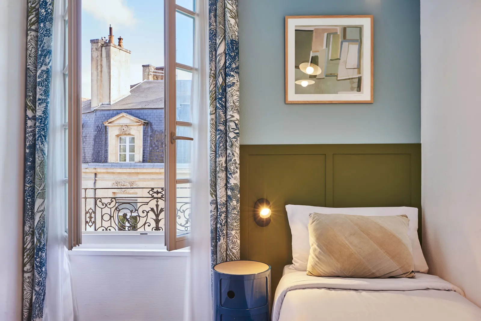 Elegant hotel bedroom featuring a bed with white linens and olive-green headboard, open window overlooking Bordeaux rooftops with wrought iron balcony, blue and white curtains, wooden-framed mirror, and black cylindrical nightstand.