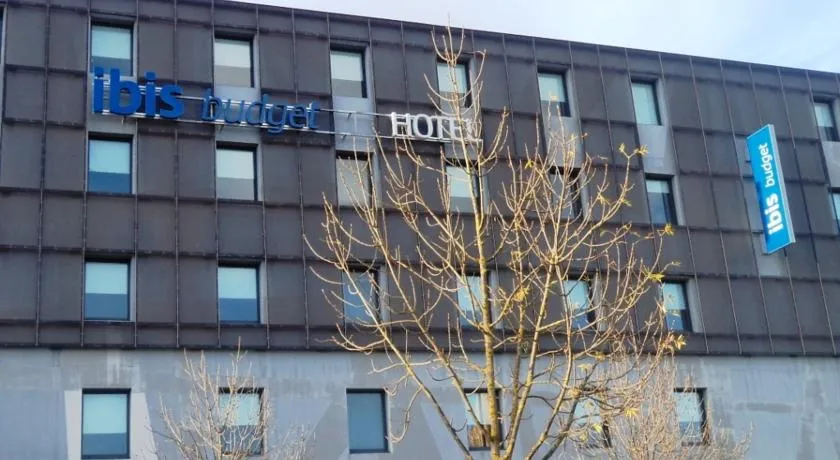 Front facade of the Ibis budget Bordeaux Centre Bastide hotel, a modern building with gray cladding, blue signage and a bare tree at the entrance