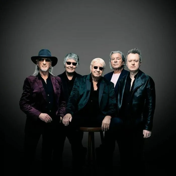 Group portrait of five Deep Purple musicians in black stage outfits and accessories, posed against a gray background.
