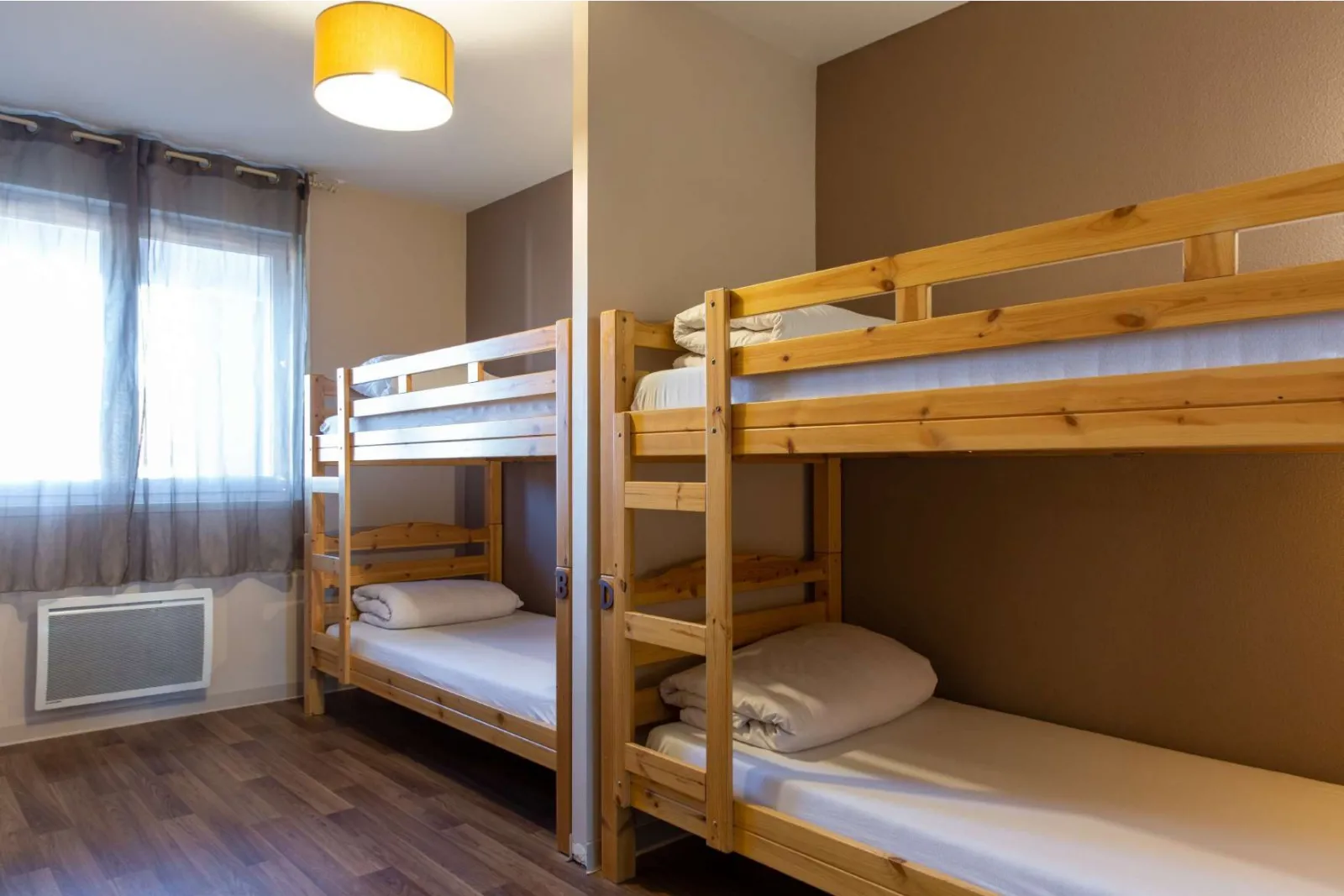 Dormitory room with two wooden bunk beds, beige walls, grey curtained window, yellow ceiling light and white heater at Bordeaux Blanquefort Youth Hostel