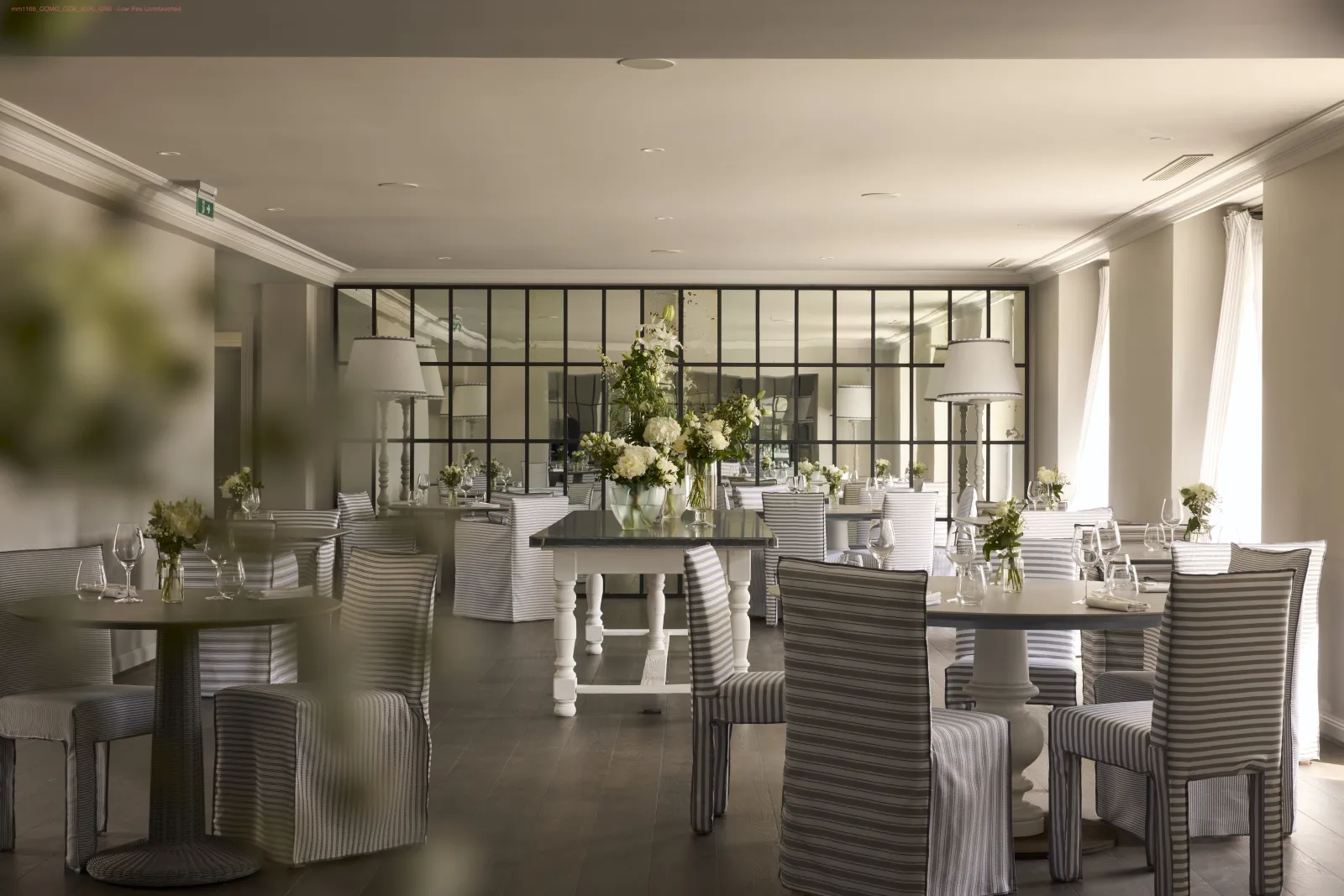 Elegant restaurant dining room at Cordeillan in Bordeaux, featuring set tables, striped upholstered chairs, large black-framed windows, modern floor lamps and white floral arrangements.