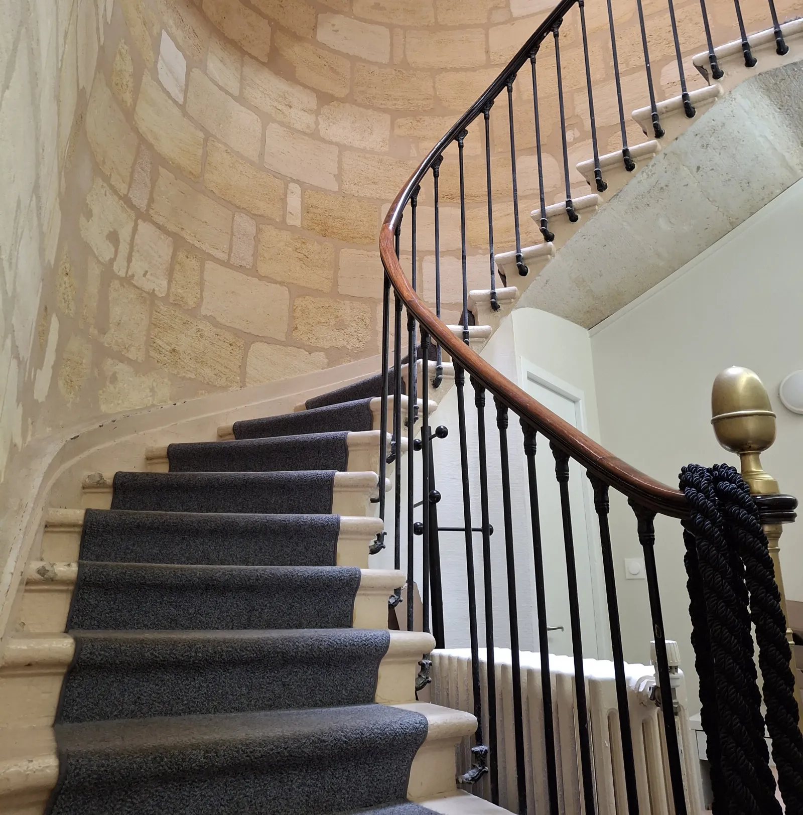 Spiral staircase with grey carpeted steps, wooden handrail and black ironwork railings, exposed stone walls, modern finish in a historic building.