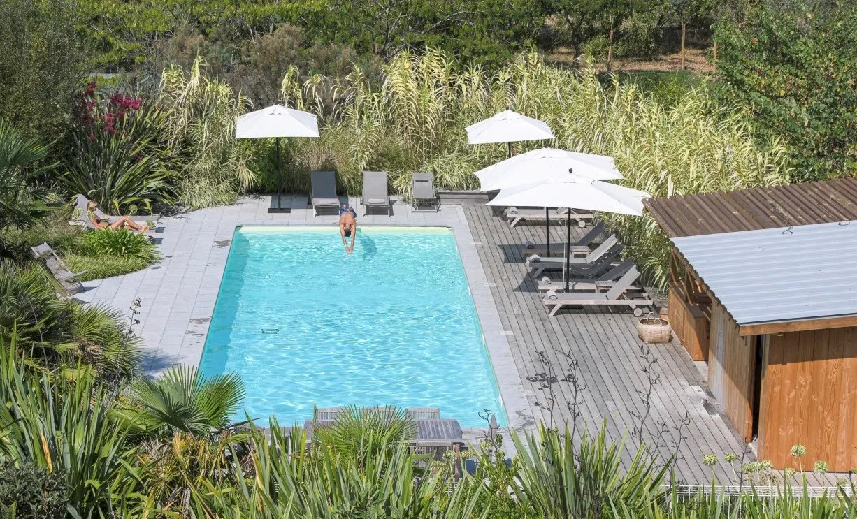 Aerial view of a turquoise rectangular swimming pool surrounded by a gray wooden deck, white lounge chairs, white umbrellas, wooden buildings, and a lush landscaped garden at Château Grattequina in Bordeaux.