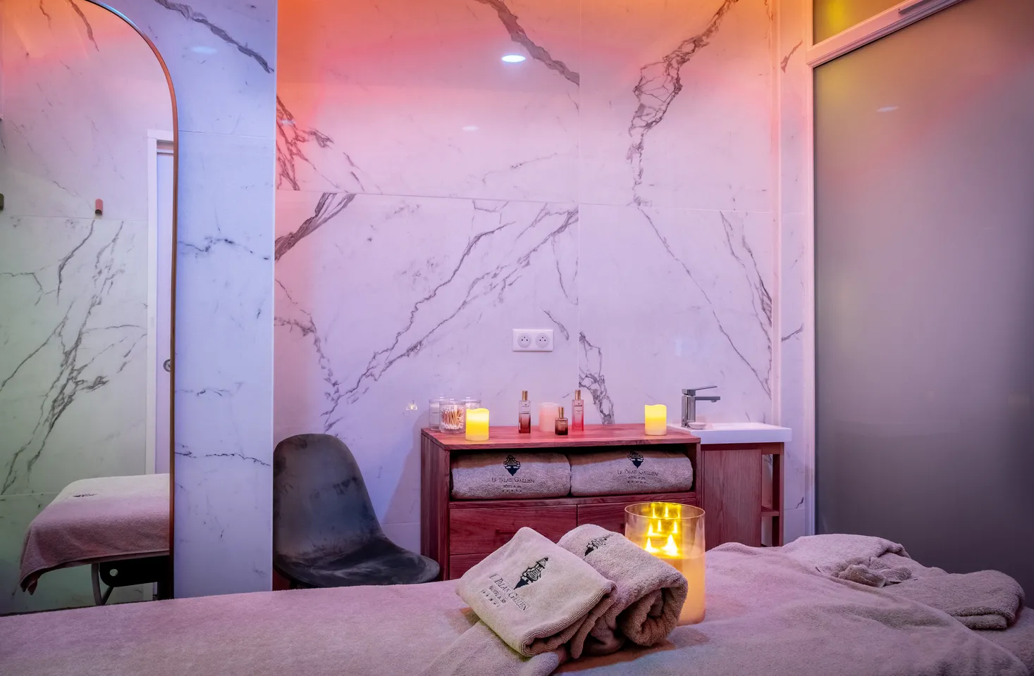 Luxurious spa treatment room featuring pink marble walls, ambient purple and pink lighting, folded white towels, yellow scented candles, and a wooden vanity with beauty products.