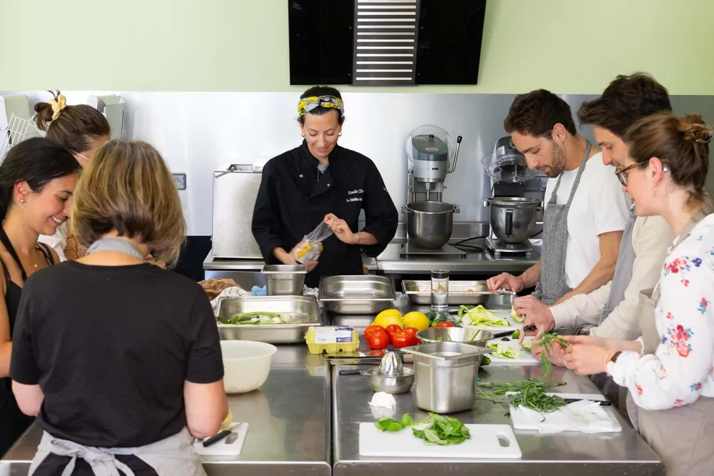 A cooking instructor in black demonstrates a culinary technique to a group of participants during a hands-on cooking class in Bordeaux. Students prepare fresh vegetables around a professional workstation equipped with electric mixers and kitchen tools.