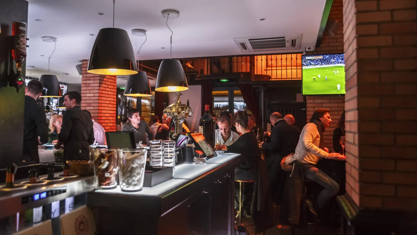 Lively interior of Bodega Bodega bar in Bordeaux with patrons at the counter, black pendant lights, exposed brick walls, and football match on screen