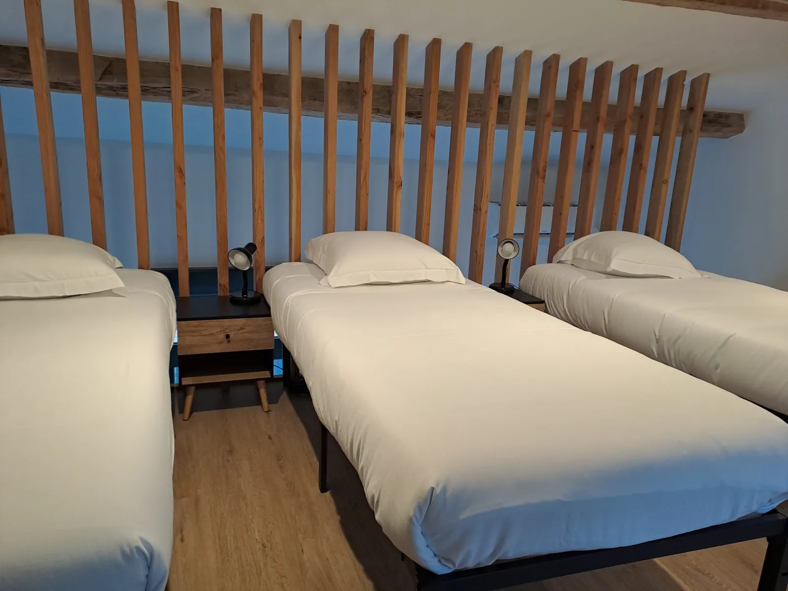 Bedroom with three single beds with white linens, wooden headboard with vertical slats, wooden floor, light blue walls, small bedside tables and reading lamps.