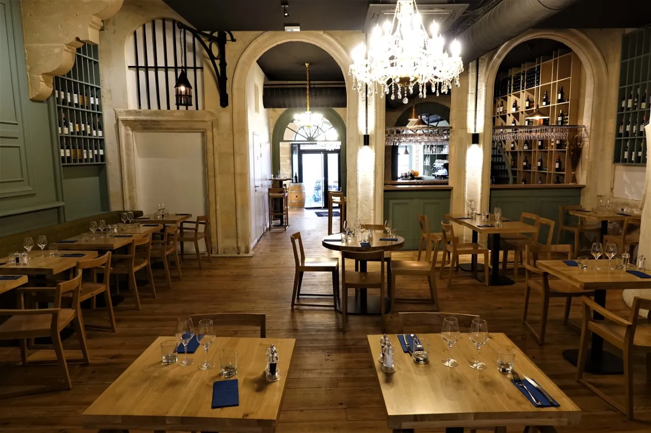 Elegant interior of Comptoir d'Etienne in Bordeaux, featuring wooden tables, rustic chairs, crystal chandeliers, architectural arches, and integrated wine storage along the walls.